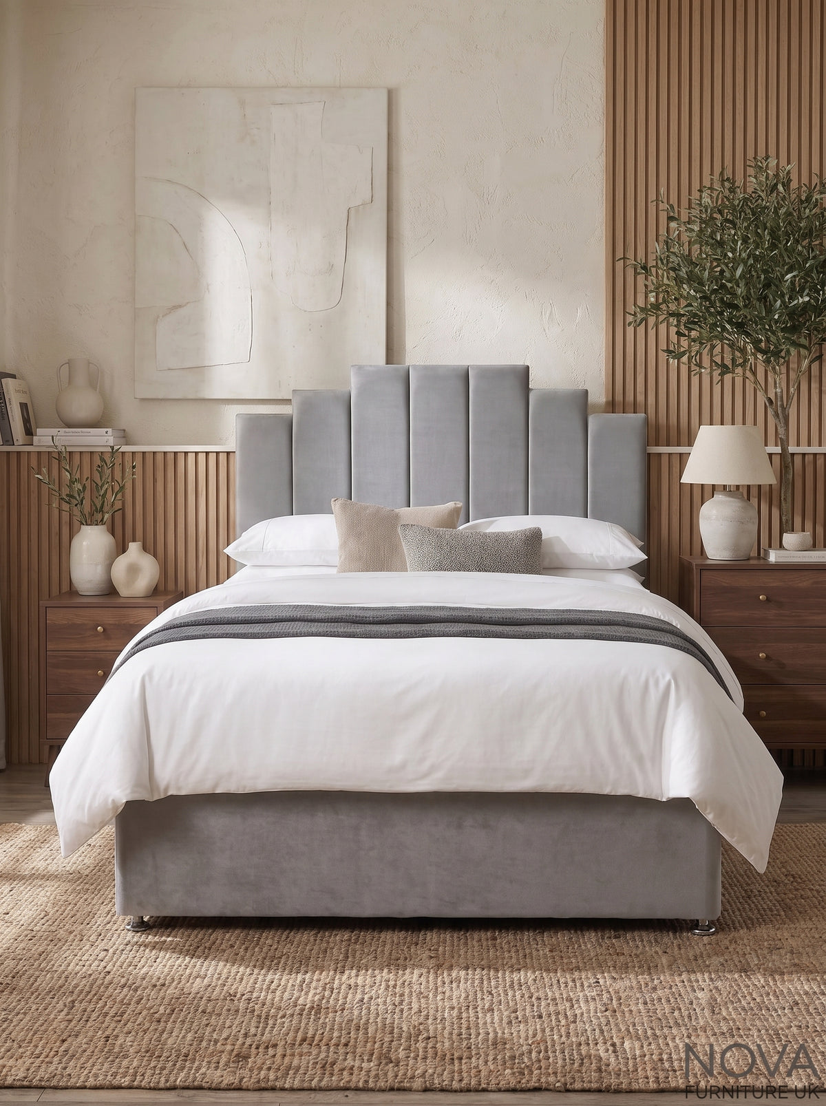 A modern bedroom scene featuring a gray upholstered bed frame with a tall segmented headboard and matching footboard, dressed in white bedding with a gray throw. The bed is styled in a warm, contemporary room with wooden nightstands, a textured beige wall, and a plant.