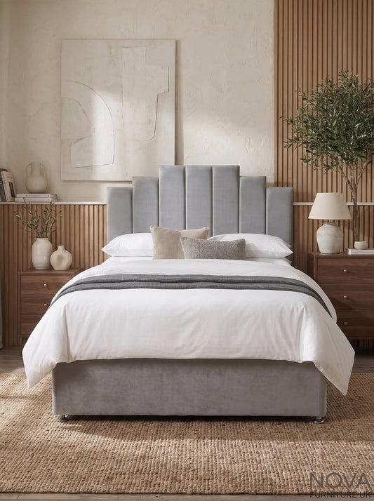A modern bedroom scene featuring a gray upholstered bed frame with a tall segmented headboard and matching footboard, dressed in white bedding with a gray throw. The bed is styled in a warm, contemporary room with wooden nightstands, a textured beige wall, and a plant.