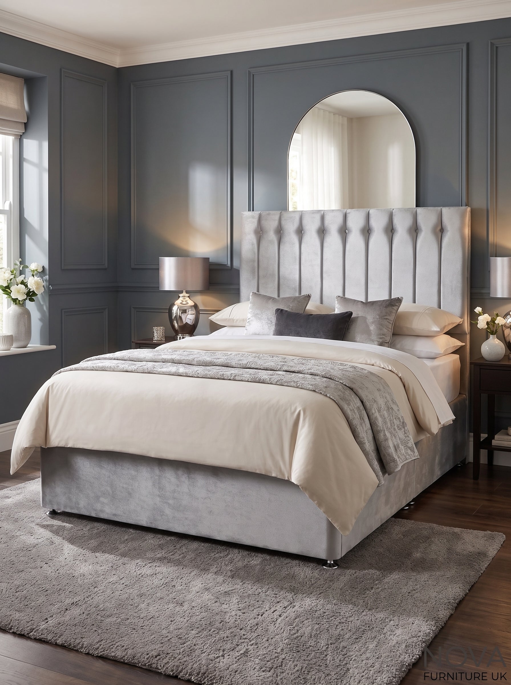 A modern bedroom featuring a bed with a tall, tufted light gray upholstered headboard and matching gray base, dressed in beige and gray bedding. The room has dark gray paneled walls, a large arched mirror, bedside tables with lamps, and a plush gray rug on dark wood floors.