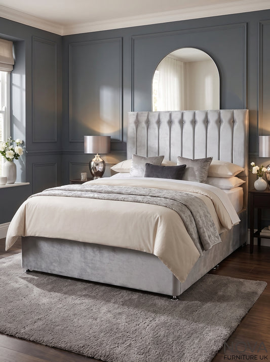 A modern bedroom featuring a bed with a tall, tufted light gray upholstered headboard and matching gray base, dressed in beige and gray bedding. The room has dark gray paneled walls, a large arched mirror, bedside tables with lamps, and a plush gray rug on dark wood floors.